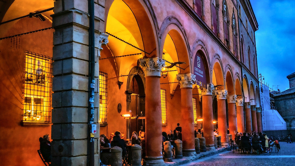People dining at dusk under the lit arches of a historic portico in Bologna