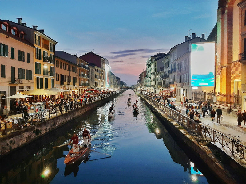 Rowing boats on the Naviglio Grande in Milan at dusk, with people at canal-side bars and restaurants