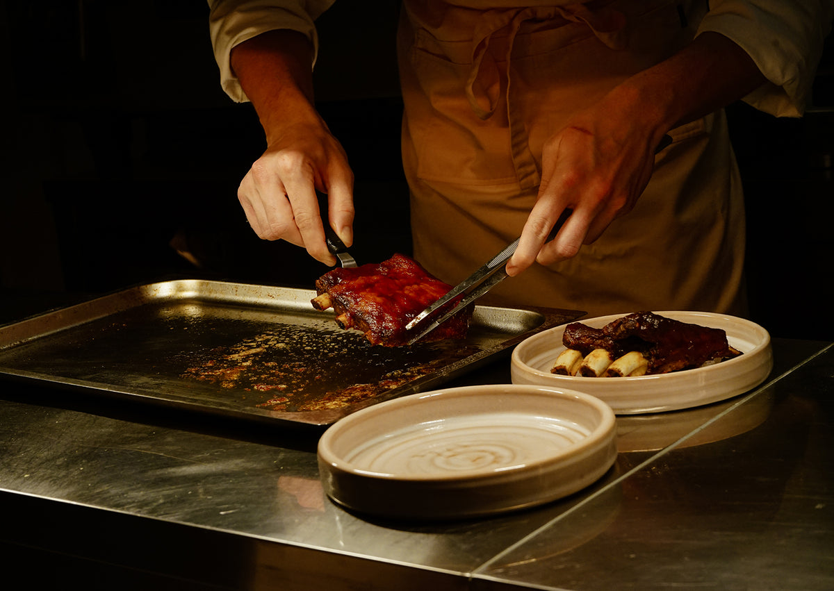 A cook plating glazed ribs from a tray in the kitchen at Ahimè