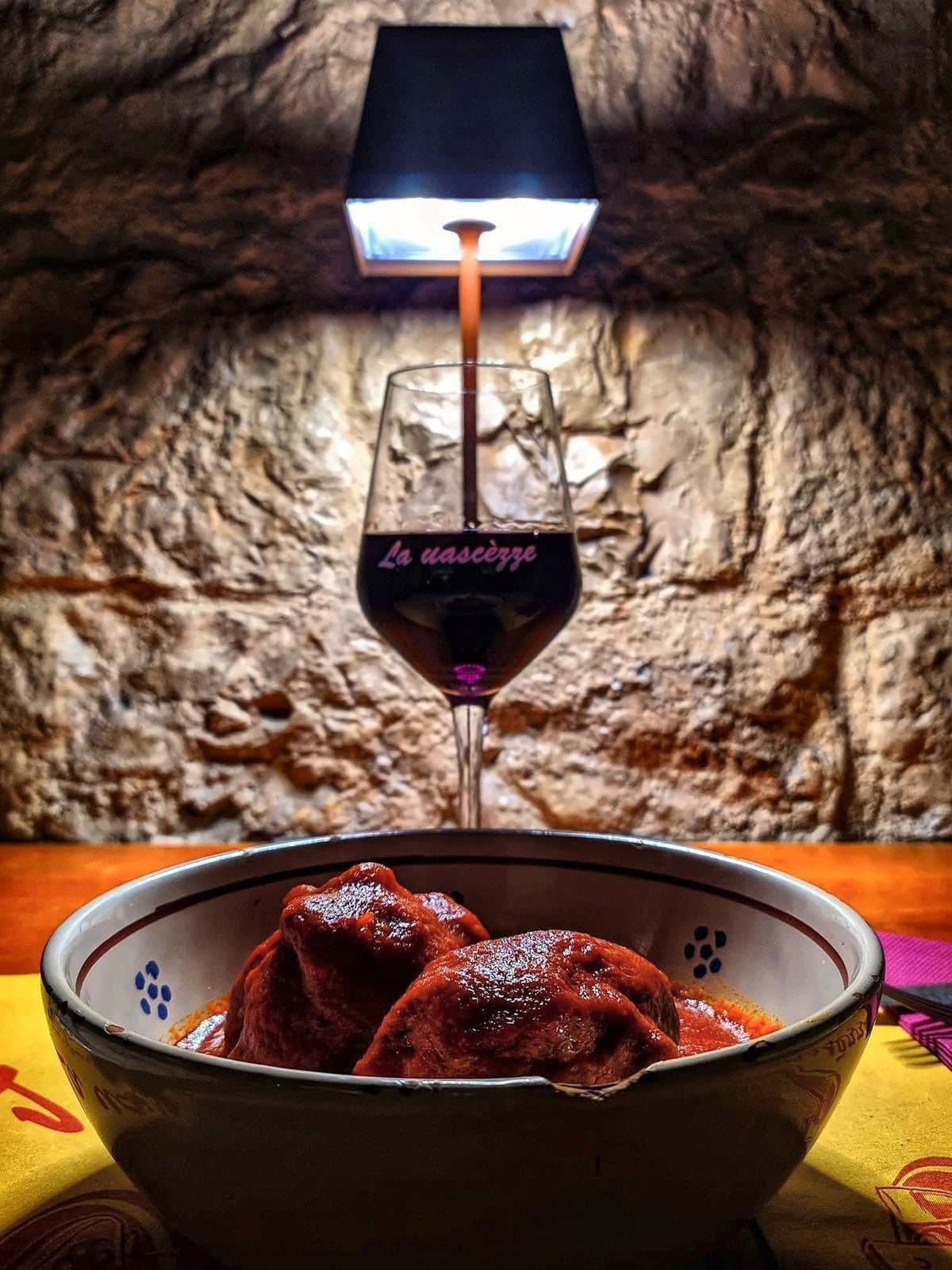 A glass of red and a bowl of meat in tomato sauce against the stone wall at La Uascezze