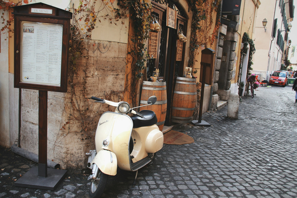 A vintage scooter parked by a trattoria menu board on a cobbled lane