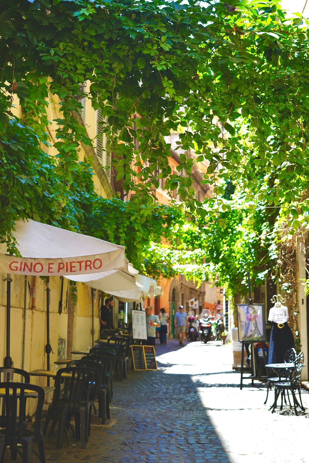 Restaurant tables under a leafy canopy in a narrow street