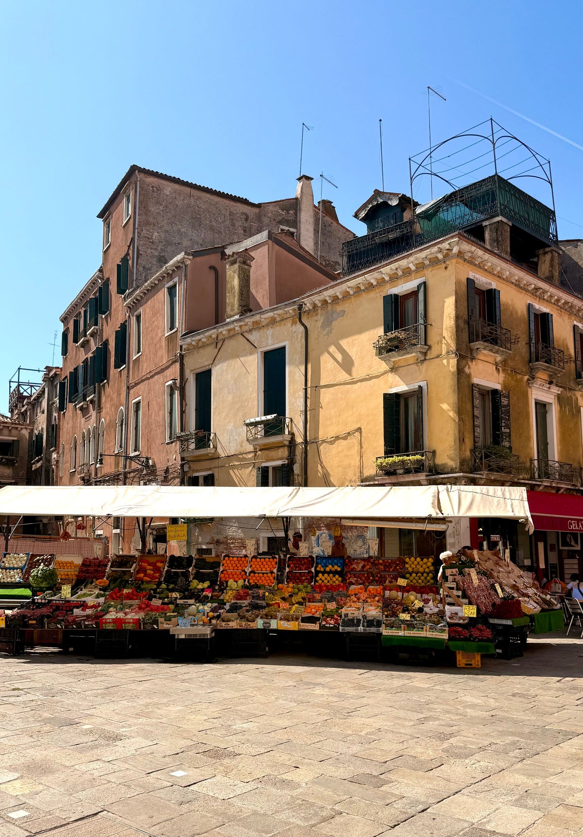 A fruit and vegetable stall under an awning in a sunny Venetian campo