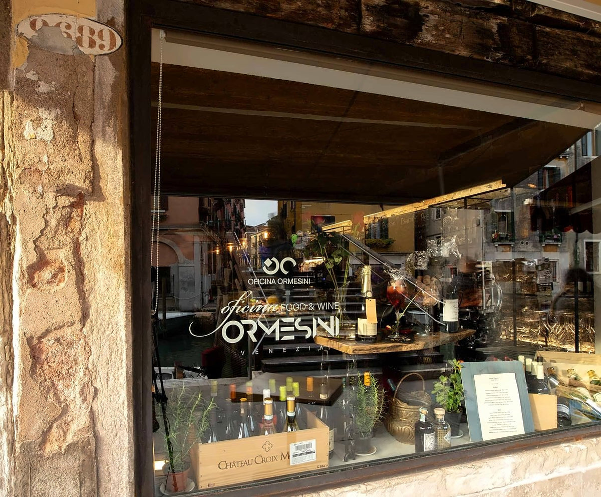 Shop window of Oficina Ormesini with wines and herbs, canal reflected in the glass